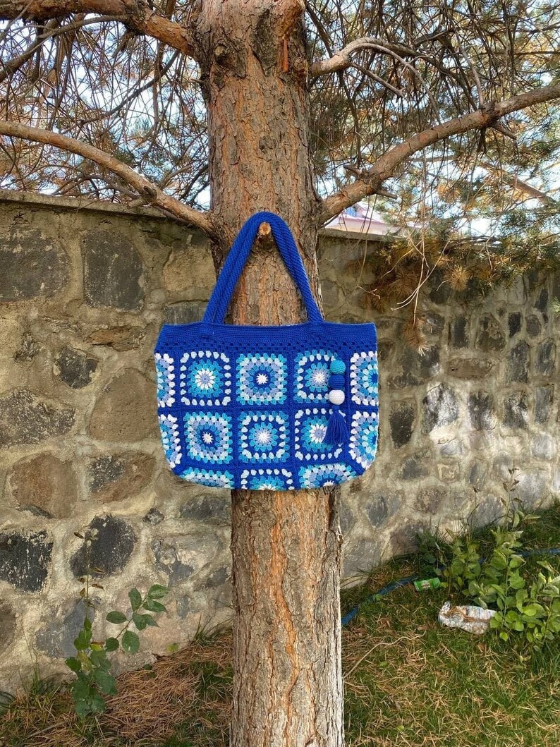 May include: A blue and white crocheted tote bag with a white pom-pom and tassel hanging from the handle. The bag is hanging on a tree trunk.