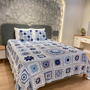 May include: A handcrafted crochet blanket with a square pattern in white, light blue, and navy blue, covering a bed. Two matching square cushions are on the bed. The blanket features floral and geometric designs. The bed is against a textured stone wall.