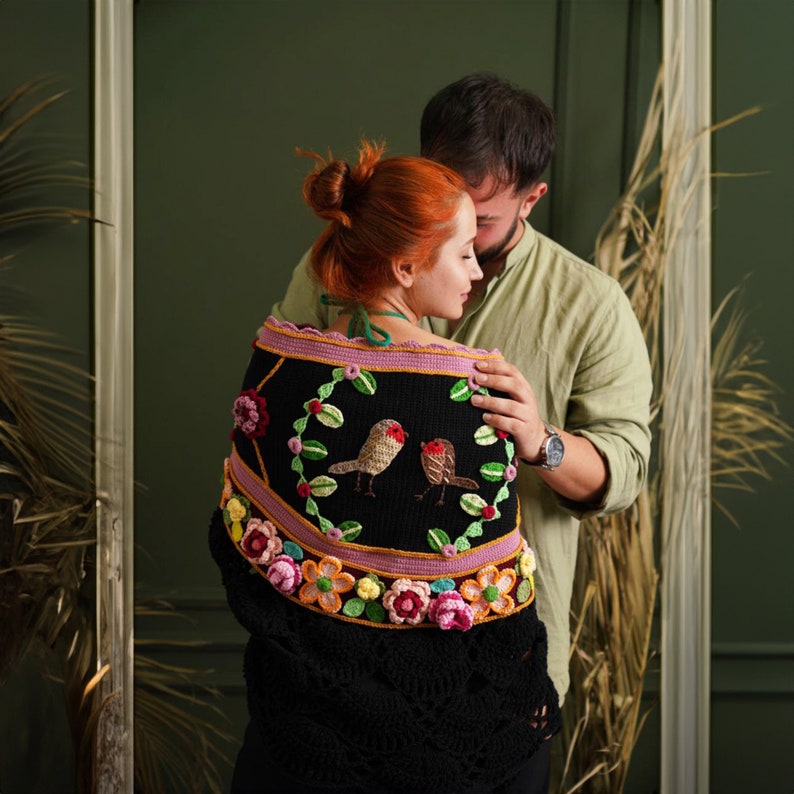 May include: A couple standing close together, the woman wearing a black crocheted shawl with colorful floral embroidery and two birds. The shawl has a pink and yellow border.