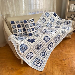 Puede incluir: Una manta de crochet blanca con patrones geométricos azules y grises. La manta está drapeada sobre un sofá blanco con dos cojines de crochet azules y grises a juego.