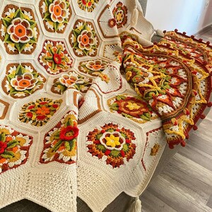 May include: A crocheted throw blanket with a white background and colorful floral patterns. The blanket has a fringe border and is draped over a chair.