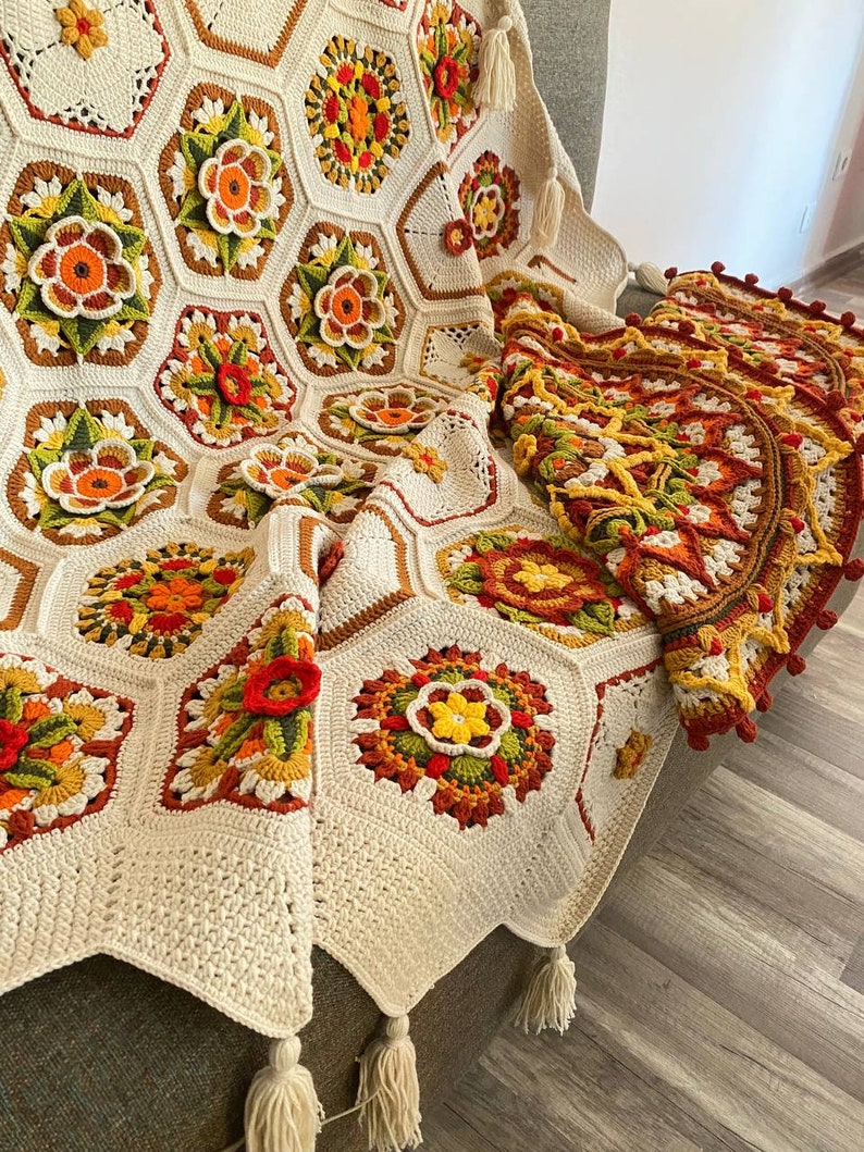 May include: A crocheted throw blanket with a white background and colorful floral patterns. The blanket has a fringe border and is draped over a chair.