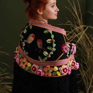 May include: A black crocheted shawl with a pink and yellow floral border and embroidered birds. The shawl is worn by a person with red hair.
