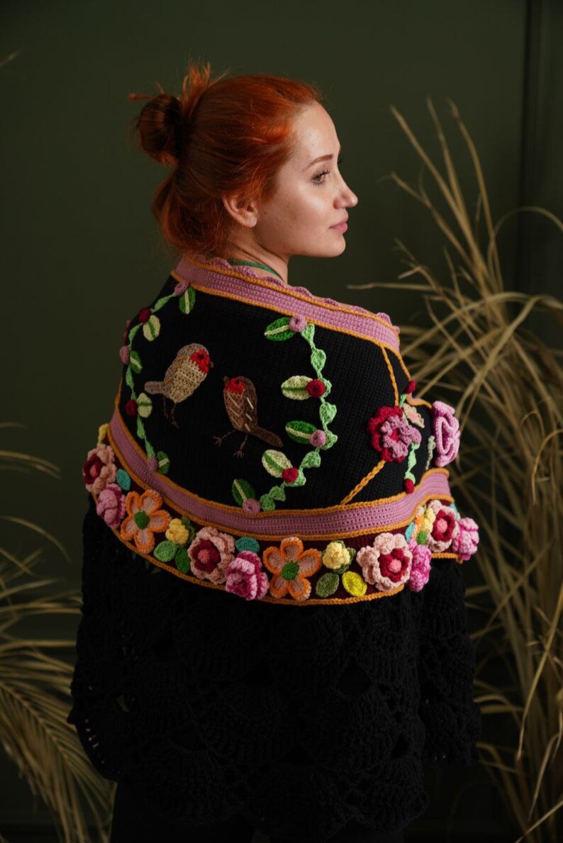 May include: A black crocheted shawl with a pink and yellow floral border and embroidered birds. The shawl is worn by a person with red hair.