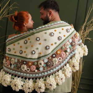 May include: A couple wearing a crocheted shawl with a floral design in shades of white, green, brown, and pink. The shawl has a fringe border and is decorated with crocheted flowers.