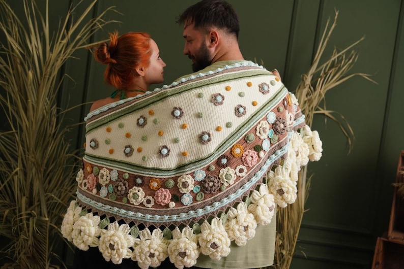 May include: A couple wearing a crocheted shawl with a floral design in shades of white, green, brown, and pink. The shawl has a fringe border and is decorated with crocheted flowers.