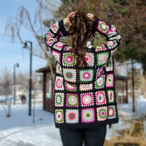 May include: A black crochet cardigan with a colorful geometric pattern. The cardigan features squares with pink, green, and white accents.
