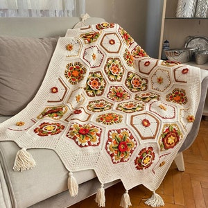 May include: A crocheted throw blanket with a white background and colorful floral designs. The blanket has a fringe border and is draped over a couch.