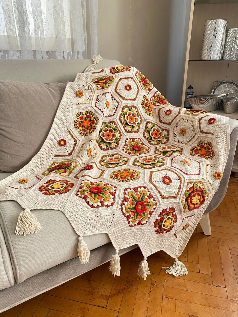 May include: A crocheted throw blanket with a white background and colorful floral designs. The blanket has a fringe border and is draped over a couch.