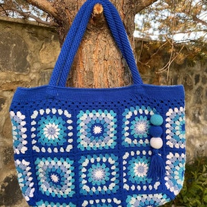 May include: A blue crocheted tote bag with a white and blue geometric pattern. The bag has a long handle and a tassel.