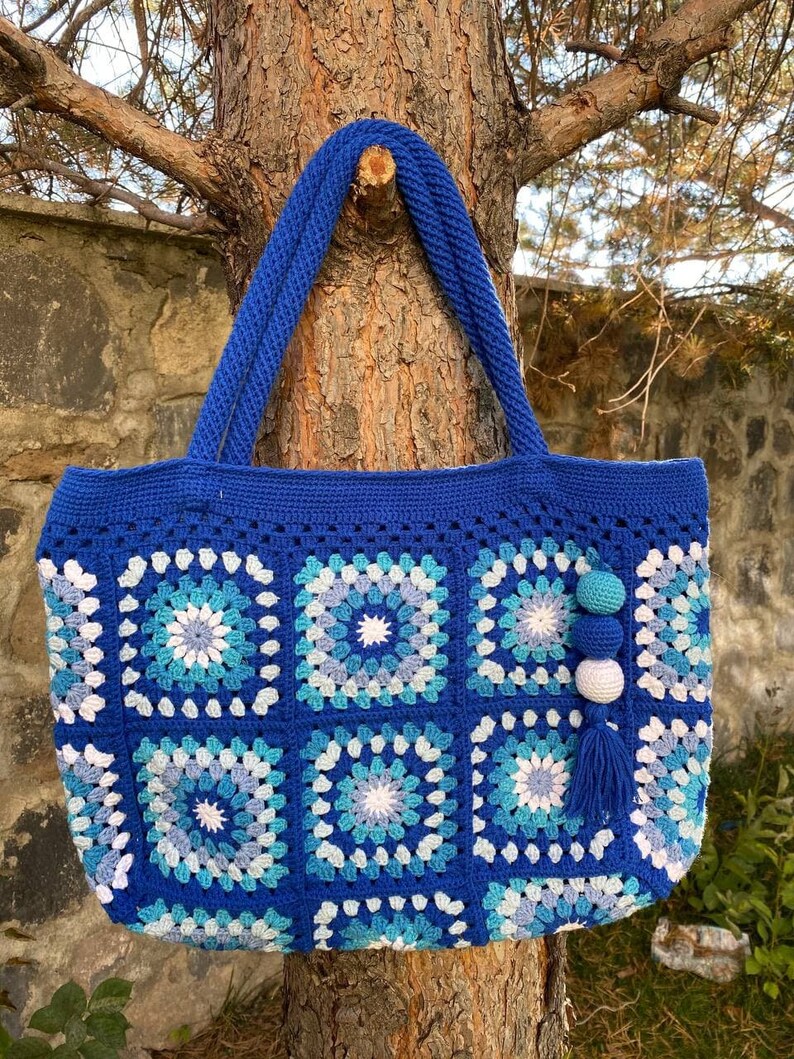 May include: A blue crocheted tote bag with a white and blue geometric pattern. The bag has a long handle and a tassel.