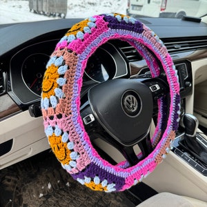 May include: A colorful crocheted steering wheel cover with a pink, purple, blue, yellow, and brown color scheme. The cover features a repeating pattern of smiley faces.