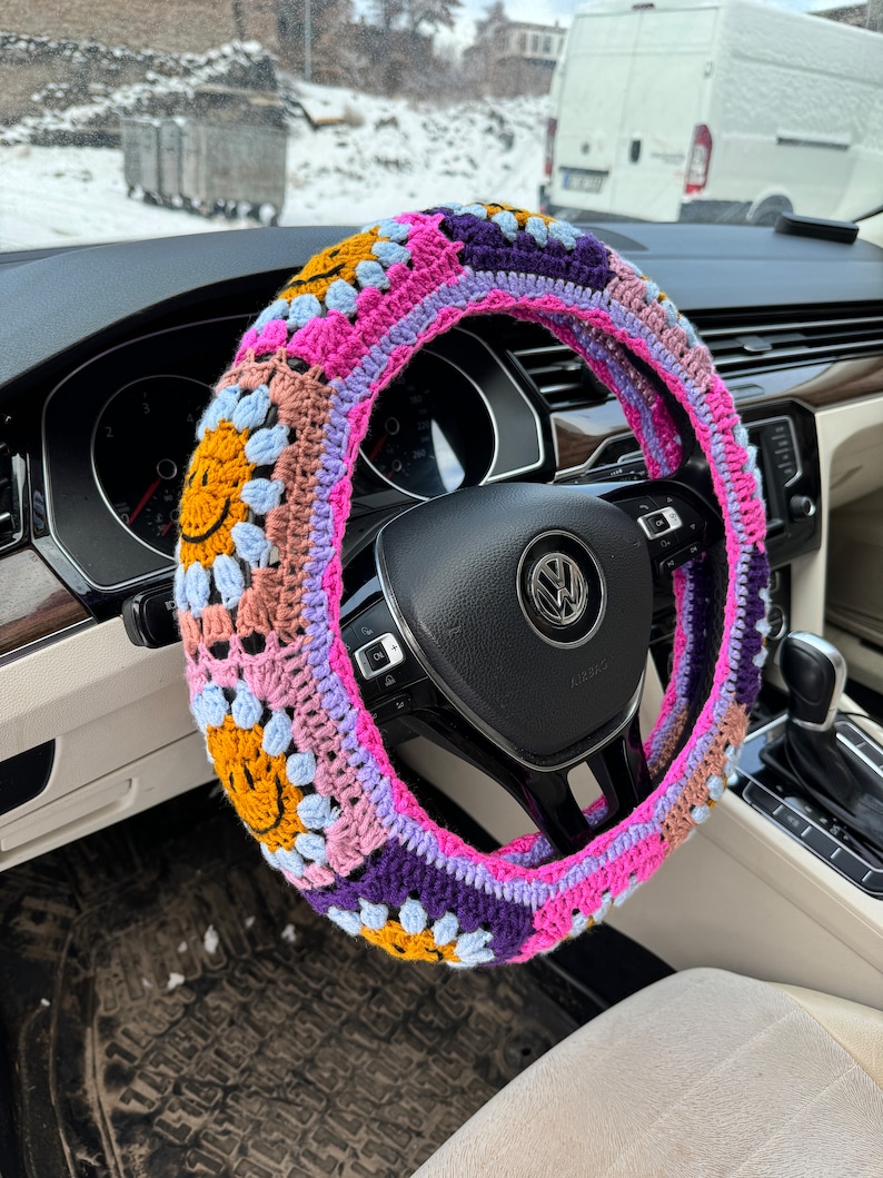 May include: A colorful crocheted steering wheel cover with a pink, purple, blue, yellow, and brown color scheme. The cover features a repeating pattern of smiley faces.
