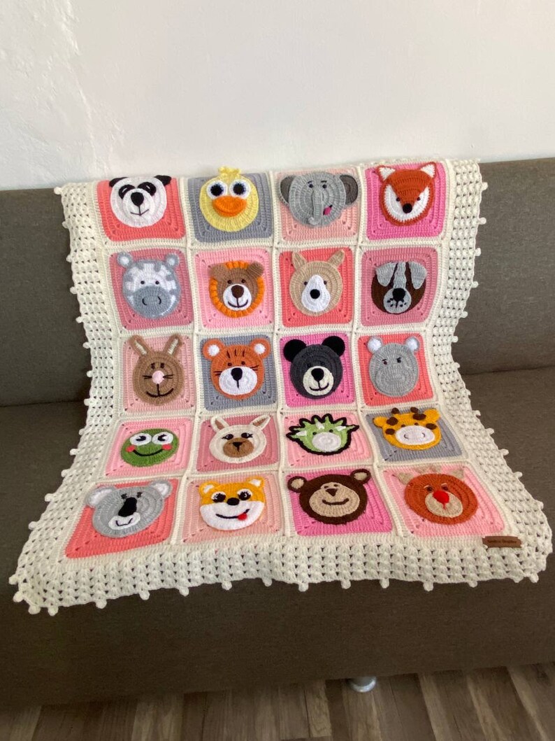 Puede incluir: Una manta de beb&eacute; de crochet con un borde blanco y cuadrados rosas con diferentes caras de animales. La manta est&aacute; hecha con hilo suave y es perfecta para la habitaci&oacute;n de un beb&eacute;.