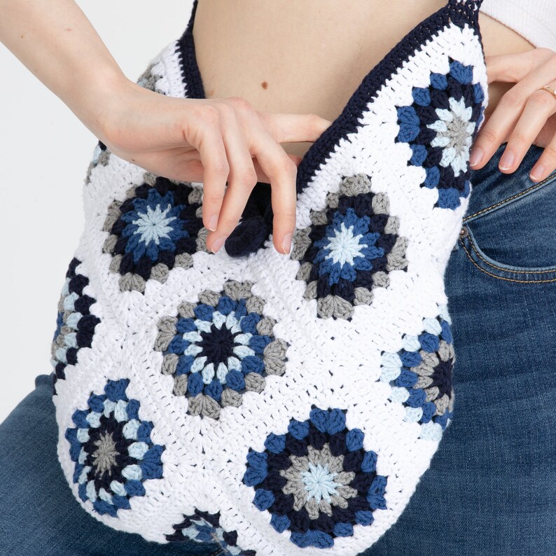 May include: A white crocheted tote bag with blue and gray granny squares. The bag has a long strap and a tassel.