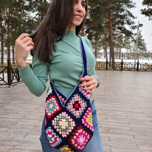 May include: A woman wearing a green mock neck shirt and blue jeans is carrying a large, colorful crocheted tote bag with a blue strap.