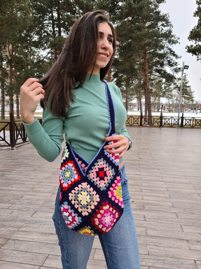 May include: A woman wearing a green mock neck shirt and blue jeans is carrying a large, colorful crocheted tote bag with a blue strap.