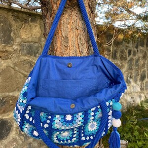 May include: A blue crocheted tote bag with a blue fabric lining and a white and blue tassel. The bag is hanging from a tree branch.