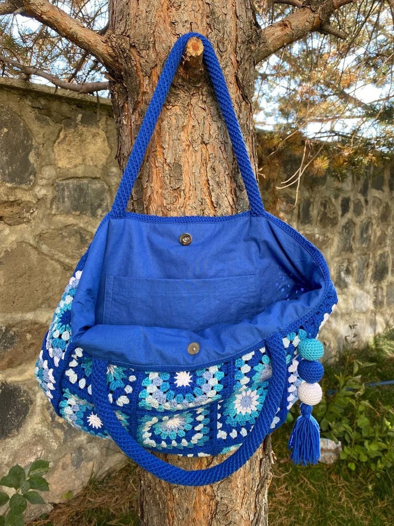 May include: A blue crocheted tote bag with a blue fabric lining and a white and blue tassel. The bag is hanging from a tree branch.