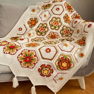 May include: A white crocheted throw blanket with a geometric pattern of hexagons in shades of brown, orange, red, and green. The blanket has tassels on the edges.