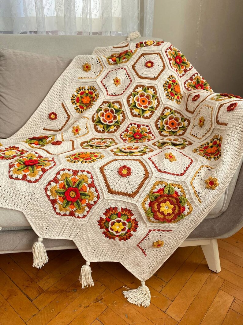May include: A white crocheted throw blanket with a geometric pattern of hexagons in shades of brown, orange, red, and green. The blanket has tassels on the edges.