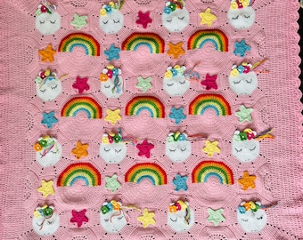 May include: A pink crocheted blanket featuring a repeating pattern of white unicorn faces with floral accents, colorful rainbows, and star shapes. The blanket has a scalloped edge and is made of soft yarn.