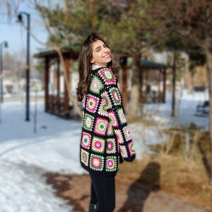 May include: A woman wearing a black and multi-colored crochet cardigan with a geometric pattern. The cardigan has pink, green, and white accents. The woman is standing in a snowy park.