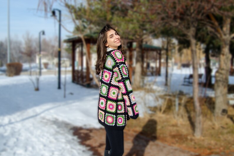 May include: A woman wearing a black and multi-colored crochet cardigan with a geometric pattern. The cardigan has pink, green, and white accents. The woman is standing in a snowy park.
