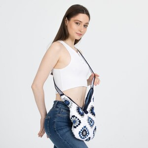 May include: A white crocheted bag with blue and white floral patterns. The bag has a long strap and is worn over the shoulder.