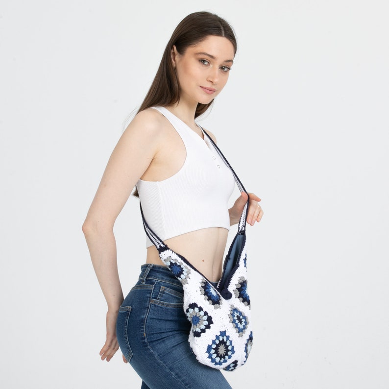 May include: A white crocheted bag with blue and white floral patterns. The bag has a long strap and is worn over the shoulder.