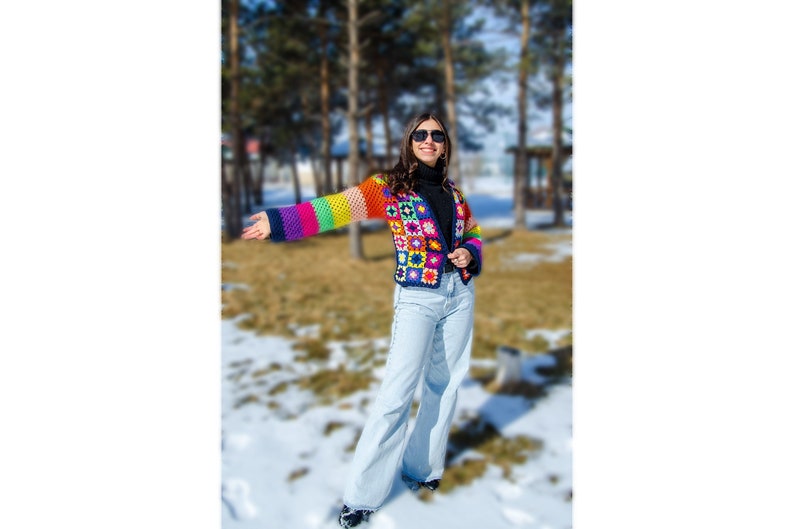 Puede incluir: Una mujer lleva una chaqueta de ganchillo de colores con un patr&oacute;n geom&eacute;trico. La chaqueta tiene rayas de arco&iacute;ris en las mangas y una base negra. Lleva pantalones vaqueros azules y gafas de sol.
