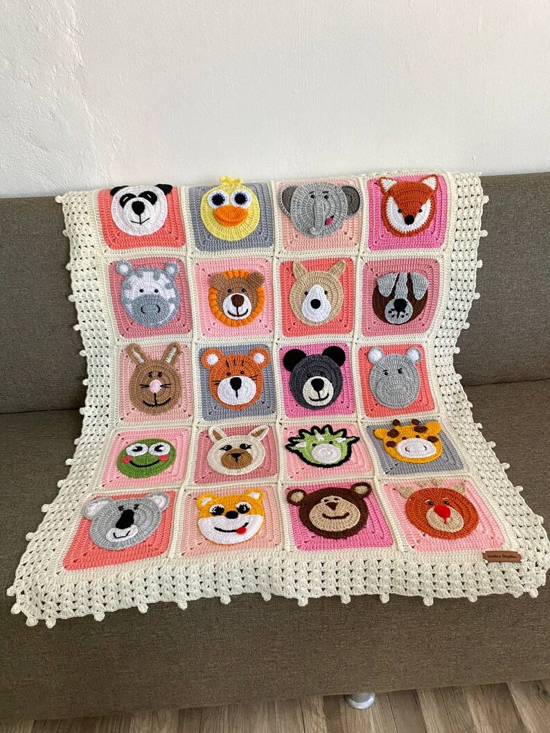 Puede incluir: Una manta de beb&eacute; de crochet con un borde rosa y blanco. La manta presenta una variedad de caras de animales, incluyendo un panda, un pato, un elefante, un zorro, una cebra, un oso, un perro, un koala, una rana, una jirafa y un mono.