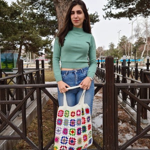 May include: A woman wearing a green turtleneck sweater and blue jeans is holding a white crocheted tote bag with colorful squares. The bag has a long strap and is hanging from her shoulder.