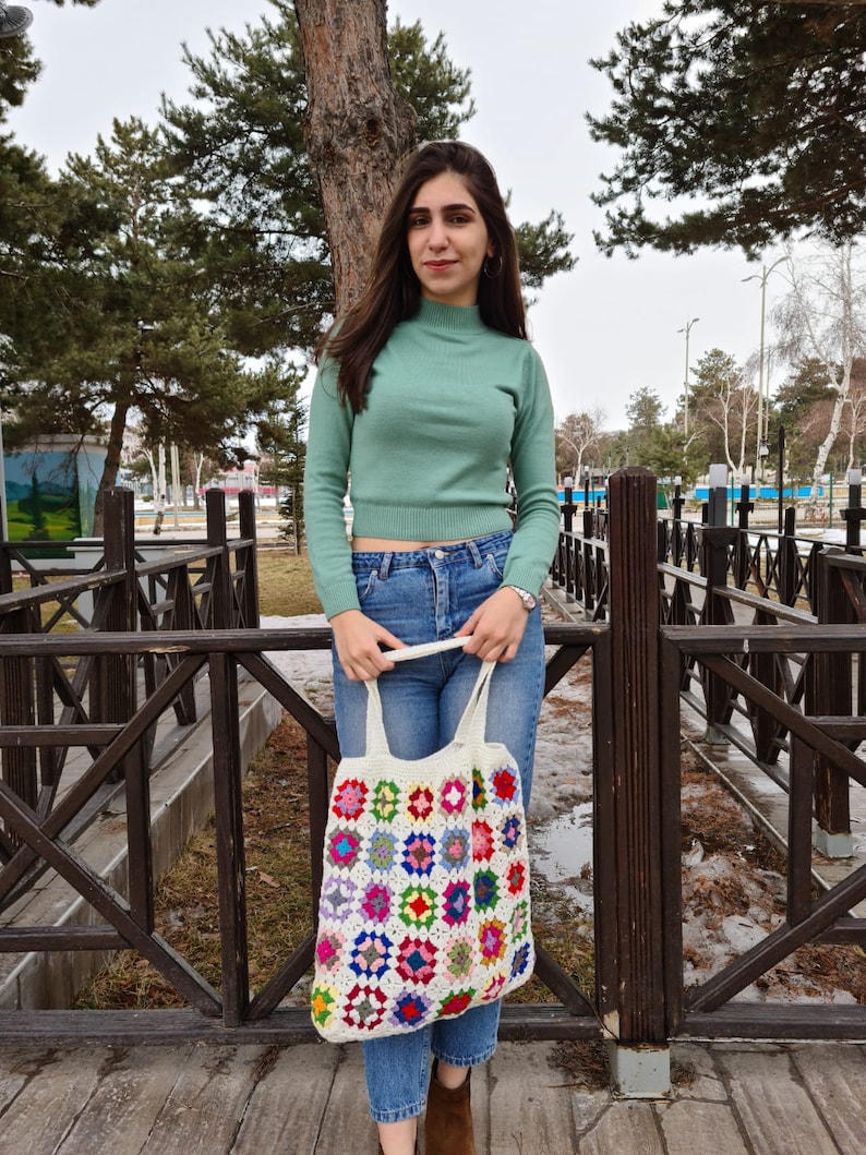 May include: A woman wearing a green turtleneck sweater and blue jeans is holding a white crocheted tote bag with colorful squares. The bag has a long strap and is hanging from her shoulder.