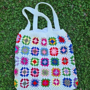 May include: A white crocheted tote bag with colorful granny square designs. The bag has two handles and is made with a white yarn.