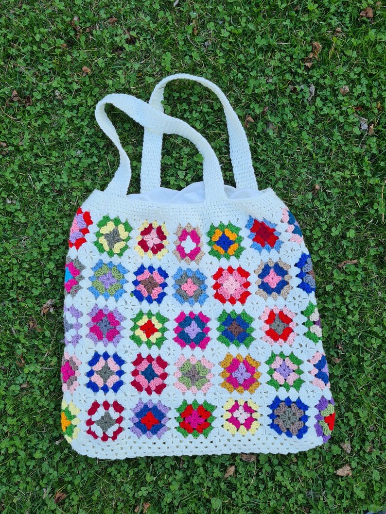 May include: A white crocheted tote bag with colorful granny square designs. The bag has two handles and is made with a white yarn.