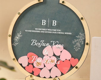 May include: A round wooden guest book with a chalkboard insert. The chalkboard has the text "BB We sincerely welcome you to our wedding and extend our cordial wishes BeiJun Xiao & Bai Yu Xi" written on it. The guest book is filled with pink and red heart-shaped wooden pieces.