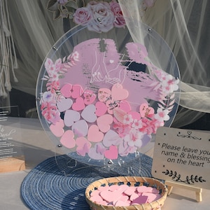 May include: A clear acrylic round wedding guest book with a pink floral design and a couple's silhouette. The guest book is filled with pink and purple heart-shaped wooden pieces for guests to sign. A sign reads "Please leave your name & blessing on the heart".