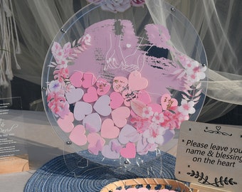 May include: A clear acrylic round wedding guest book with a pink floral design and a couple's silhouette. The guest book is filled with pink and purple heart-shaped wooden pieces for guests to sign. A sign reads "Please leave your name & blessing on the heart".