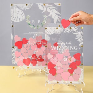 Puede incluir: Un libro de visitas de boda de acrílico transparente con un diseño floral y el texto "Welcome to the Wedding of Bei Jun Xiao & Bai Yu Xi 28|07|2022". Los invitados pueden dejar sus buenos deseos colocando tarjetas en forma de corazón rosa y rojo dentro de la caja.