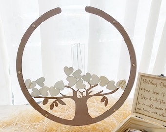 May include: A wooden wedding guest book in the shape of a tree with a circular frame. The tree has many small heart-shaped pieces that guests can write on and drop into the frame. The frame is made of wood and has a light brown finish. The sign reads "Wedding Guest Please Sign A Heart Drop it into the frame so we will never forget our loved ones who came."