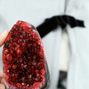 May include: A dark red crystal geode with a black base. The crystal structure is visible, with many small, sparkling facets. The geode is held in a hand, with a light-coloured background.