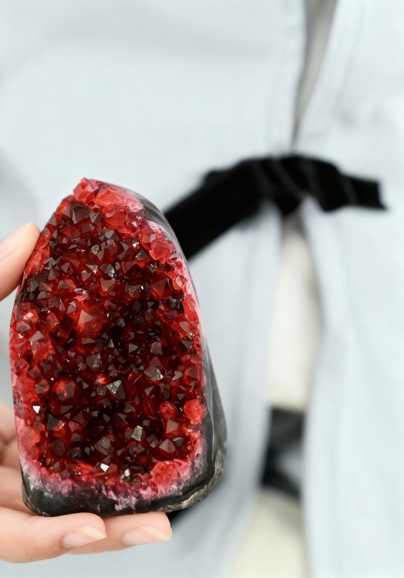 May include: A dark red crystal geode with a black base. The crystal structure is visible, with many small, sparkling facets. The geode is held in a hand, with a light-coloured background.