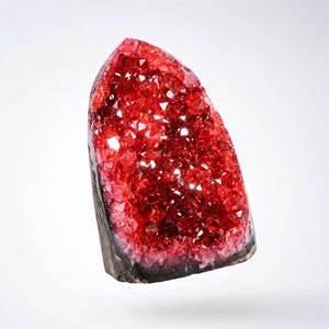 K&ouml;nnte beinhalten: Eine leuchtend rote Kristallgeode mit einer dunkelgrauen Basis. Das Innere ist mit funkelnden, kleinen Kristallformationen gef&uuml;llt. Die Geode hat eine nat&uuml;rliche, organische Form, und die Kristalle fangen das Licht ein und erzeugen einen schillernden Effekt.