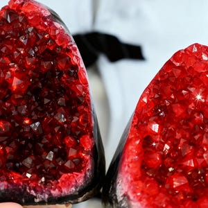 May include: Two halves of a geode with a vibrant red crystal interior. The crystals sparkle, contrasting with the dark, rough exterior. The geode is held up against a white background.