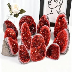 May include: A display of red crystal geodes with glittering interiors. The geodes are of various sizes and shapes, arranged on a white table. A framed artwork and a vase with a white rose are in the background.