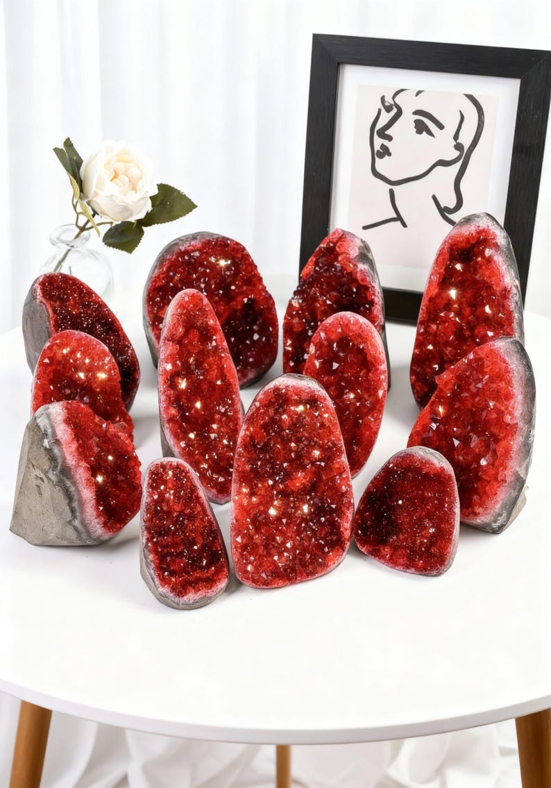 May include: A display of red crystal geodes with glittering interiors. The geodes are of various sizes and shapes, arranged on a white table. A framed artwork and a vase with a white rose are in the background.