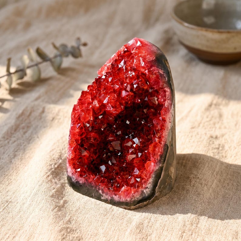 K&ouml;nnte beinhalten: Eine auff&auml;llige rote Kristallgeode mit dunkelgrauer Au&szlig;enseite. Das Innere ist mit funkelnden, tiefroten Kristallen gef&uuml;llt. Die Geode wird auf einer beigefarbenen Stoffoberfl&auml;che mit einer kleinen Keramikschale im Hintergrund pr&auml;sentiert.