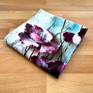 May include: A square piece of fabric with a floral print. The fabric features a large, pink and purple flower with a white center. The background is a light blue and gray watercolor wash.