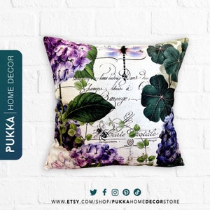 Puede incluir: Una almohada cuadrada con fondo blanco y un diseño floral. El diseño presenta hortensias moradas, hojas verdes, una libélula y una abeja. El texto "Carte Postale" está escrito en cursiva en la almohada.